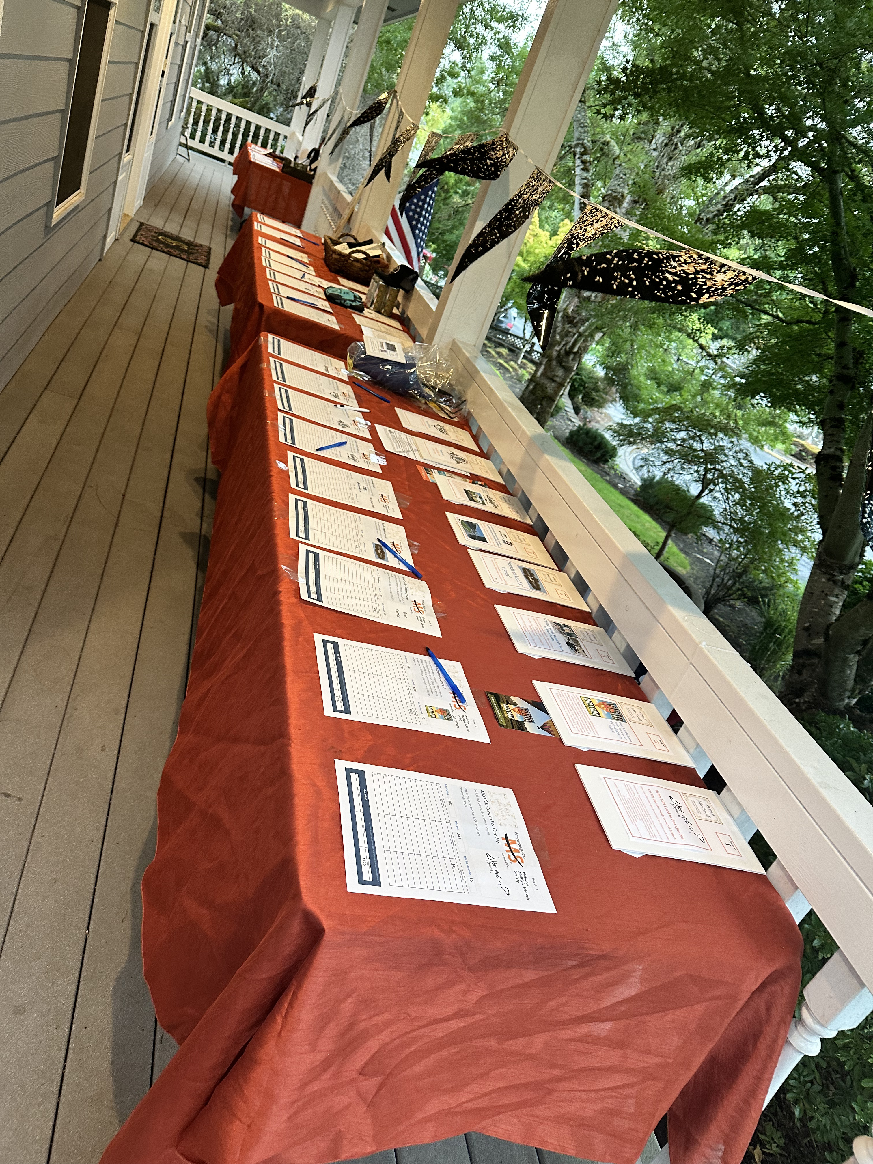 3 Long tables covered in orange tablecloths with Auction bid sheets and signs on top. The tables are on a front porch of a home.
