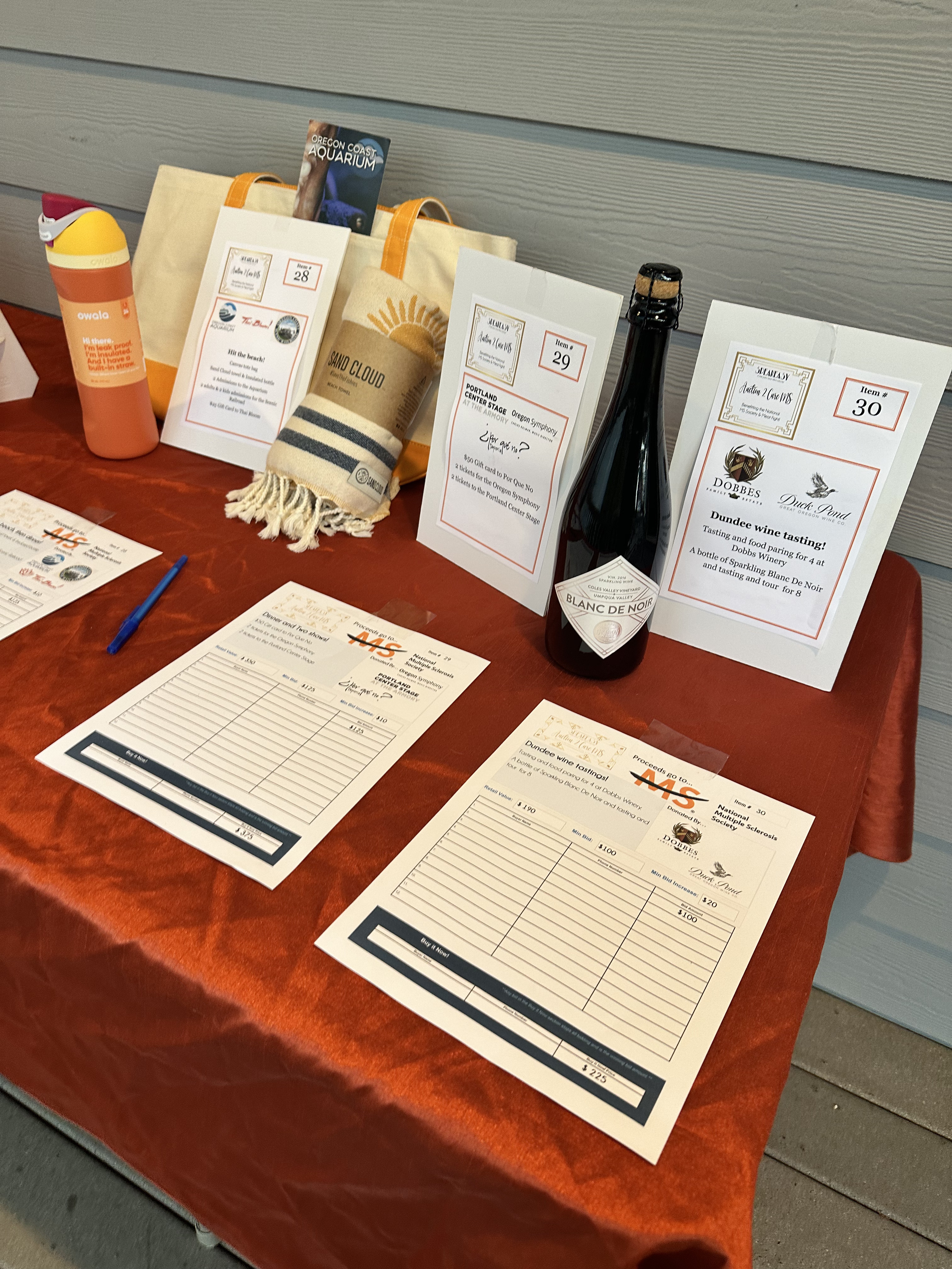 Auction item from Duck Pond winery in Dundee with a beach bag, blanket and water cup on top of the table with bid sheets in the front.