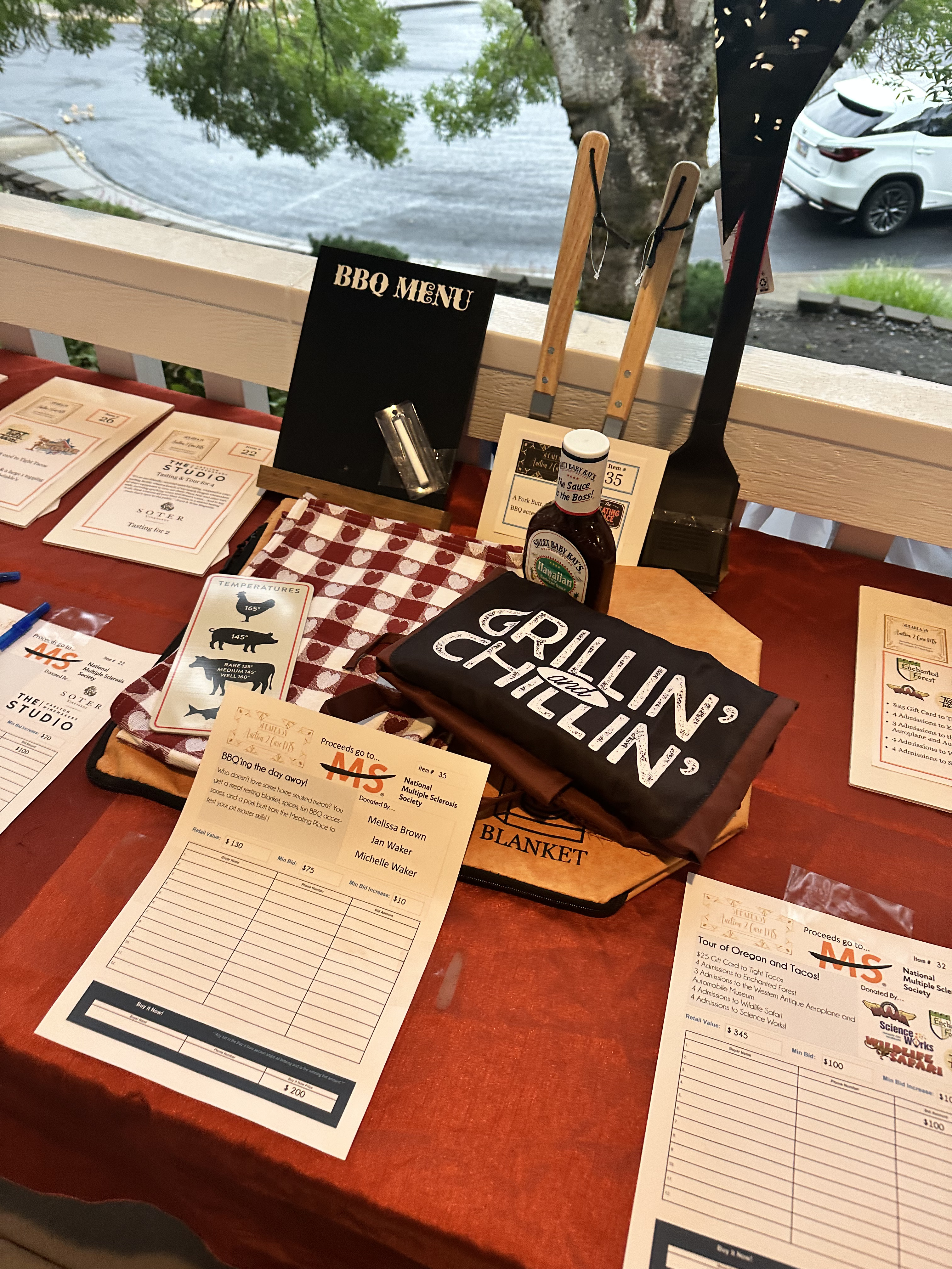 A grill theme auction item, with an apron that says grillin' and chillin', BBQ sauce, tools and a menu board sitting on the table with a bid sheet in front.
